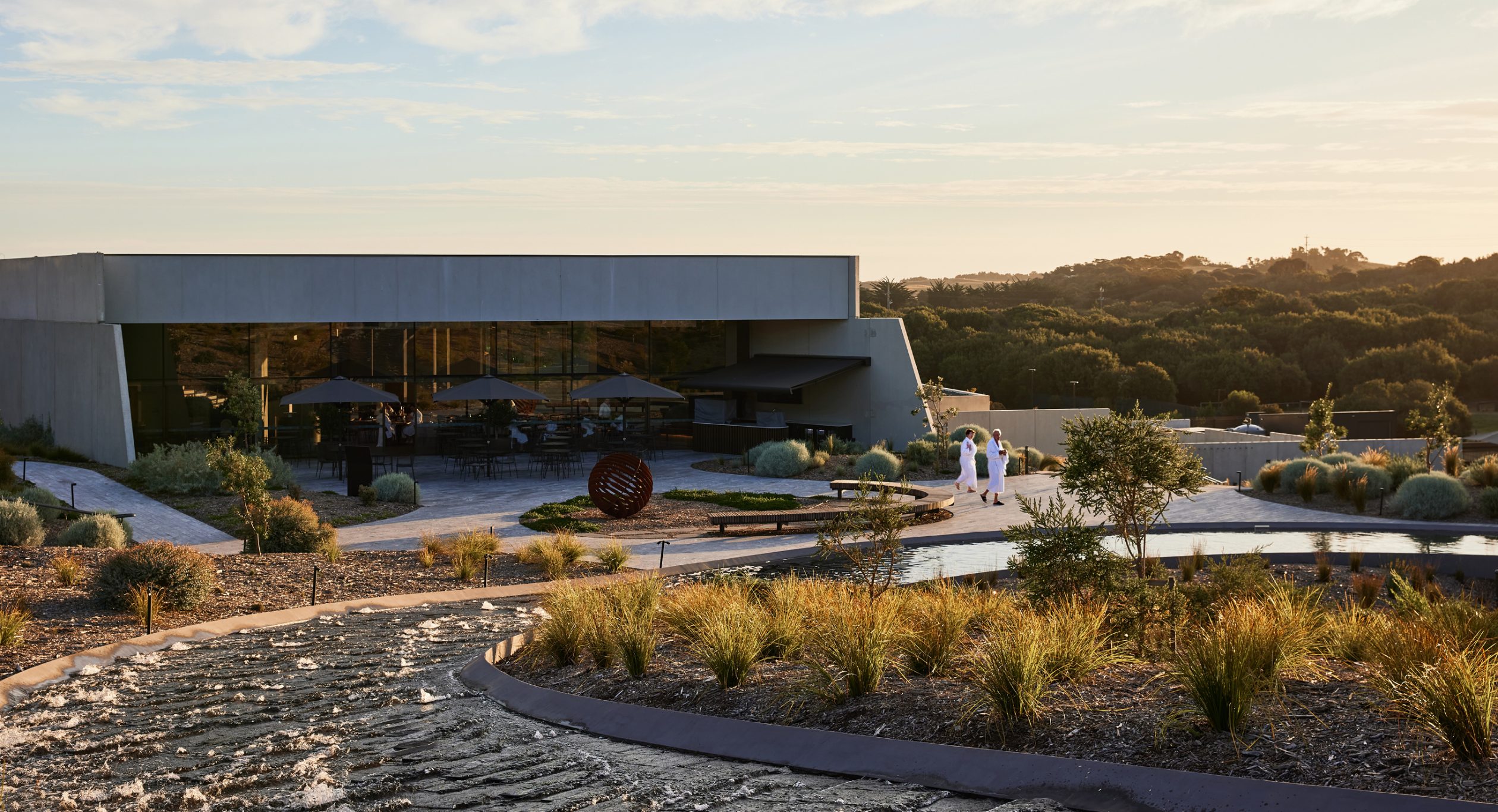 A photo of Alba Thermal Springs looking back towards the main building across the central curved water feature called "The Rapids" - two bathers in white bathrobes walk past a sculpture made from corten steel with the restaurant "Thyme" in the background and small wayfinding signs at the key decision points - in the foreground and background is a landscape of endemic plants ﻿that was designed by the landscape architect Mala.