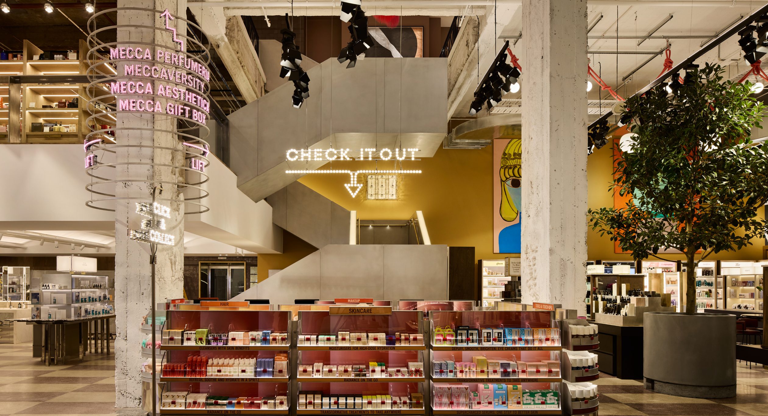 An interior view of a multi-level retail environment with an led array sign that reads "Check it out" above a cashier desk, beautiful prodcut display cases, carefully arrange bespoke tables, large tree, walls covered in artworks and a large pink halo directional sign encircles a structural column.